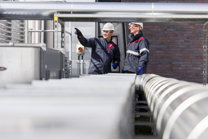 Two men standing on a roof wearing overalls, goggles and hard hats, one is pointing 